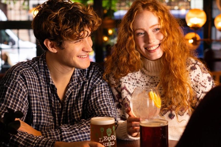 A smiling young couple enjoying drinks at a cozy pub. The man holds a pint of beer while the woman holds a cocktail garnished with lemon slices.