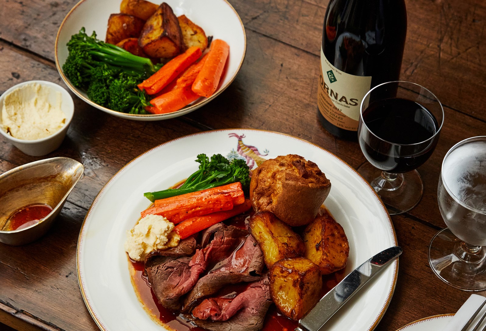 British Sunday roast served on a decorative plate, featuring slices of roast beef with gravy, roasted potatoes, glazed carrots, tenderstem broccoli, a Yorkshire pudding, and horseradish sauce. Accompanied by a bowl of extra vegetables, a glass of red wine, and a bottle of wine on a wooden table.