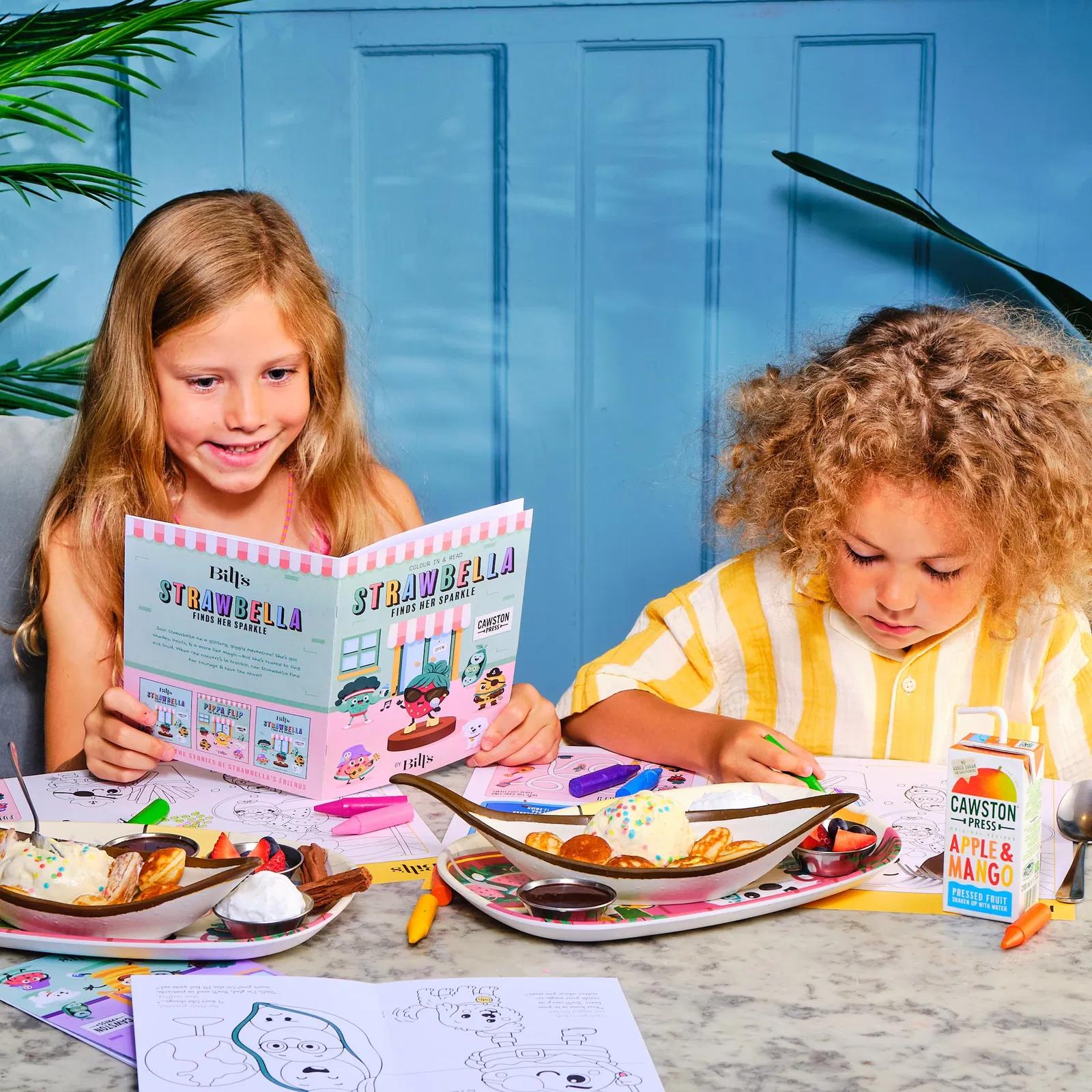 two children enjoying a meal and using activity packs reading and colouring in.